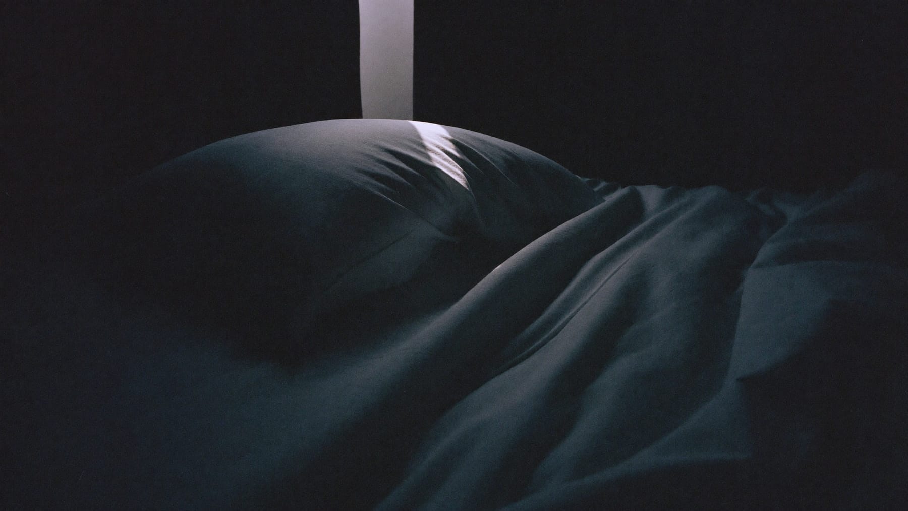 Almofada e edredão vazios num quarto quase às escuras, com uma réstia de luz a atravessar a cama