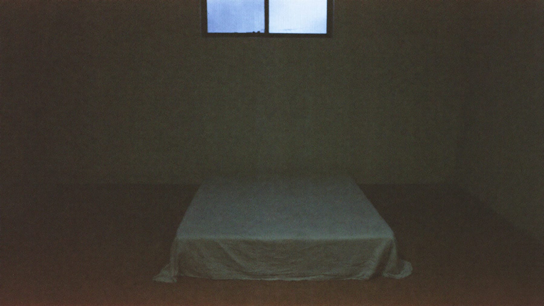 Quarto despojado com uma cama estreita e um cobertor esticado, iluminado apenas por uma janela alta